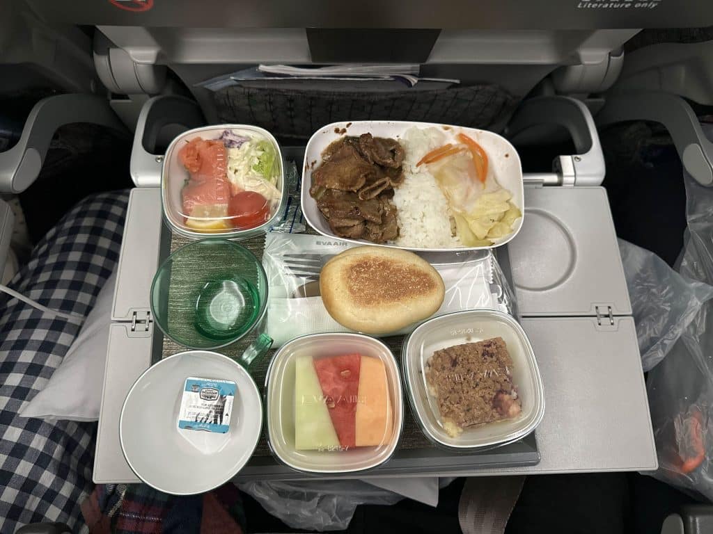 EVA Air economy class in-flight meal tray with braised beef and rice, mixed salad, fresh fruit, bread roll, and dessert bar.